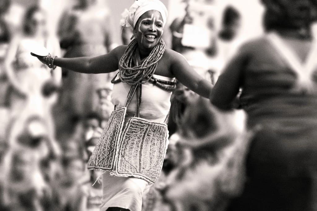 A dancer in beads and a head wrap mid-step, in black and white.